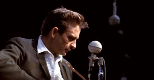 A man in a suit playing an acoustic guitar, reminiscent of Johnny Cash.
