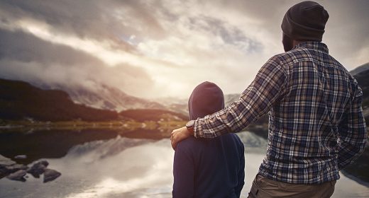 A man and his son standing by a lake with mountains in the background.