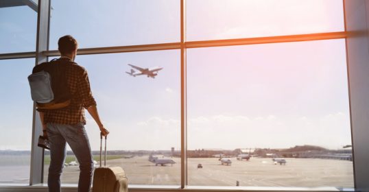 A man with a backpack looking out of an airport window, possibly booking a flight.