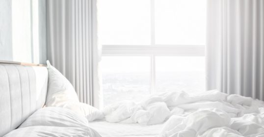 A white bed with freshly made white sheets and pillows in front of a window.