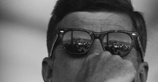 JFK's sunglasses are compellingly reflected in a mirror.