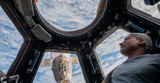 A man looking out of the window of a space station, experiencing the vastness of space.