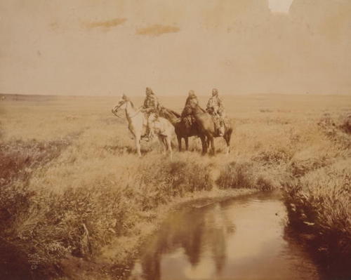Three native American warriors on horseback near a stream.