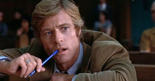 A man wearing corduroy sits in a courtroom with a pen in his hand.