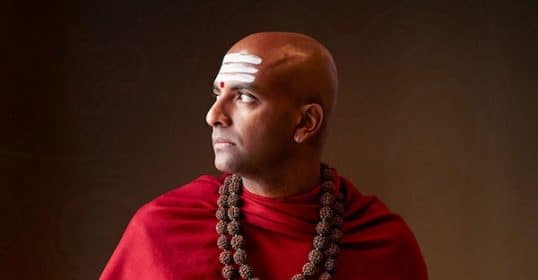 A man in a red robe with a beaded necklace exudes power and unwavering focus.