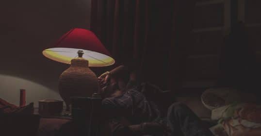 A man laying on a couch with a lamp beside him, ready to sleep.