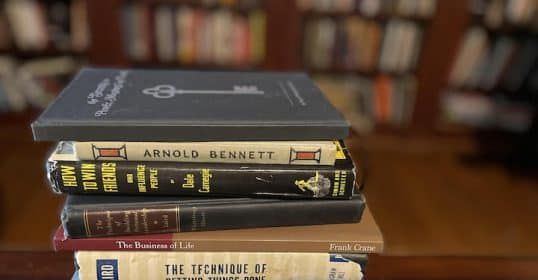 A stack of vintage books on a table in front of a bookcase.