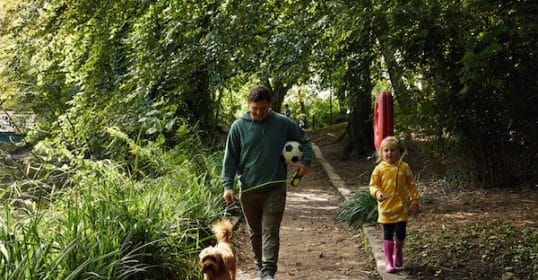 Man walking a dog on a leash with a young child carrying an analog soccer ball on a wooded path.