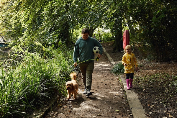 Man walking a dog on a leash with a young child carrying an analog soccer ball on a wooded path.