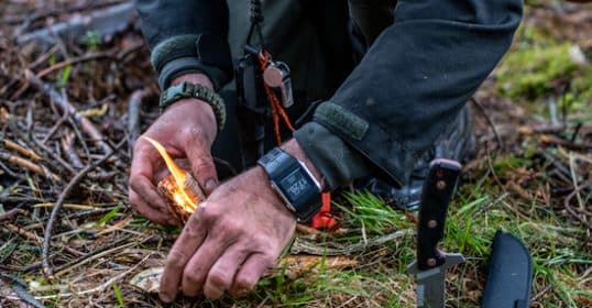 Person starting a fire with a flint in the wilderness for short-term survival, with a knife on the ground nearby.