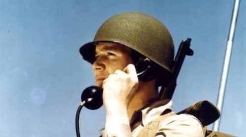 Military personnel communicating via radio handset against a clear blue sky, using the Military Phonetic Alphabet for clarity in transmission.