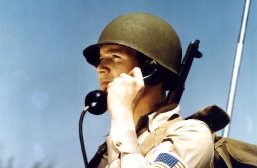 Military personnel communicating via radio handset against a clear blue sky, using the Military Phonetic Alphabet for clarity in transmission.