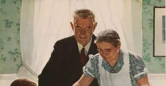 Elderly couple presenting a large turkey to a family gathered around a dinner table in their Thanksgiving outfits.