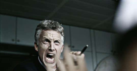 A man in a suit appears to be speaking passionately or engaging in stress management, holding a pen and gesturing with his hand.
