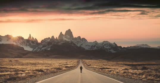 A lone person walking down a straight road towards jagged mountains at sunset, contemplating 