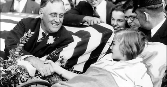 A man in a suit sitting beside a child in a wheelchair, both smiling and interacting warmly, surrounded by spectators. The child is a survivor of polio.