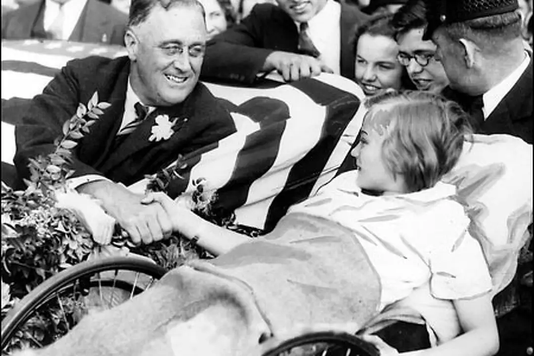 A man in a suit sitting beside a child in a wheelchair, both smiling and interacting warmly, surrounded by spectators. The child is a survivor of polio.
