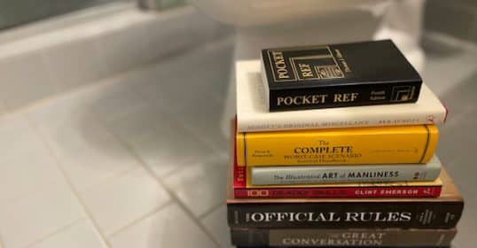 A stack of assorted books on a surface, including titles on survival skills, manliness, and cultural literacy, serves as perfect toilet reading material.