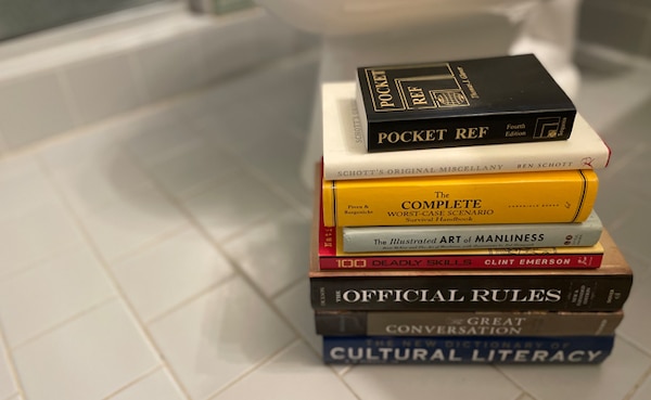 A stack of assorted books on a surface, including titles on survival skills, manliness, and cultural literacy, serves as perfect toilet reading material.