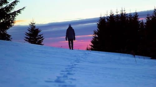 Person walking on a snow-covered slope at dusk with a colorful sky in the background, inspired by 