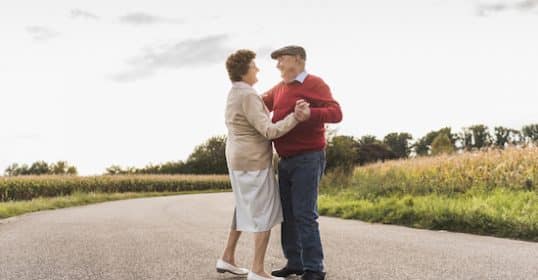 Elderly couple sharing a moment of happiness as they hold hands and gaze into each other's eyes in the middle of a deserted road with grasslands in the background.
