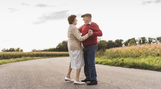 Elderly couple sharing a moment of happiness as they hold hands and gaze into each other's eyes in the middle of a deserted road with grasslands in the background.