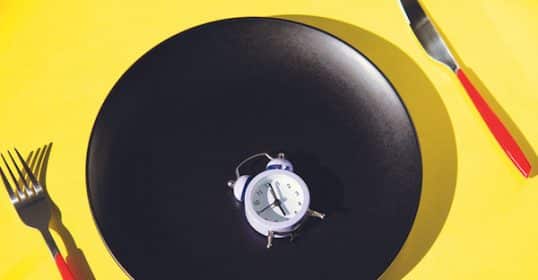 A small alarm clock centered on a black plate with a fork on the left and a knife on the right, all against a yellow background, symbolizing Extended Fasting.