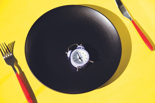 A small alarm clock centered on a black plate with a fork on the left and a knife on the right, all against a yellow background, symbolizing Extended Fasting.