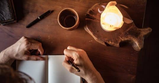 Person journaling techniques in a notebook by candlelight with a cup of coffee nearby.