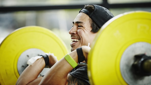 A person smiling while lifting a barbell with yellow weights at the gym and listening to a "Heal the Mind" podcast.