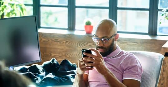 A focused man using a smartphone at a desk with a computer monitor and personal items nearby, unknowingly sabotaging his social life.