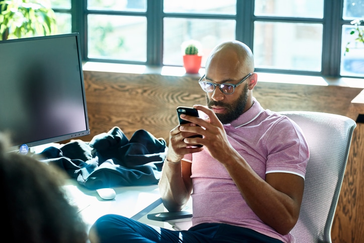 A focused man using a smartphone at a desk with a computer monitor and personal items nearby, unknowingly sabotaging his social life.