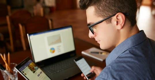 Man in glasses working on a laptop while holding a smartphone and a tablet, epitomizing the shrinking attention span.
