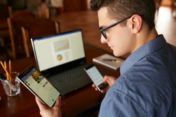 Man in glasses working on a laptop while holding a smartphone and a tablet, epitomizing the shrinking attention span.