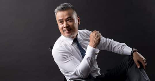 Mature man in a white shirt and black trousers sitting on a chair against a gray background, smiling and looking off-camera, exuding authority.