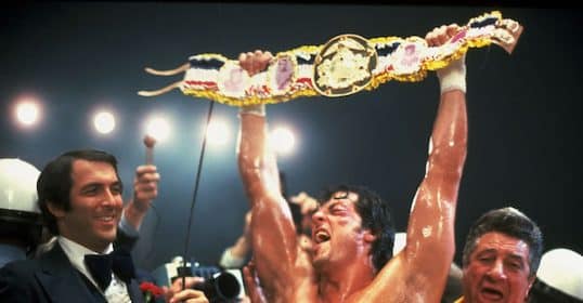 Victorious boxer, reminiscent of the Rocky movies, raises championship belt aloft as onlookers celebrate his triumph.