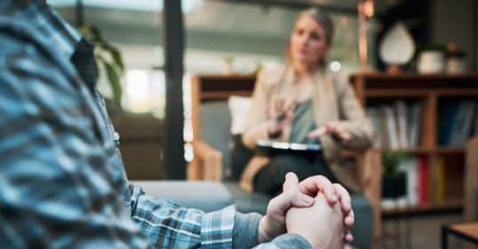 Man in a plaid shirt sitting and listening intently to a woman therapist who is taking notes during a psychology counseling session in a cozy office environment.