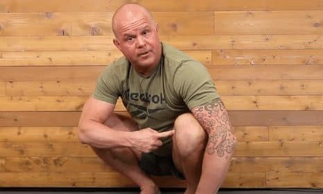 Man squatting and pointing to his tattooed arm in a wooden-paneled room, embodying the essence of an Agile Human Being.