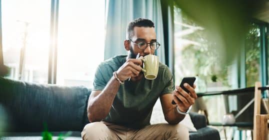 Man drinking coffee and listening to Podcast #888 on his smartphone in a sunlit room with paperwork on the table.