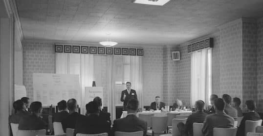 A black and white photo of a Toastmasters meeting with a speaker presenting in front of seated attendees.