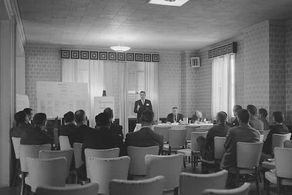 A black and white photo of a Toastmasters meeting with a speaker presenting in front of seated attendees.