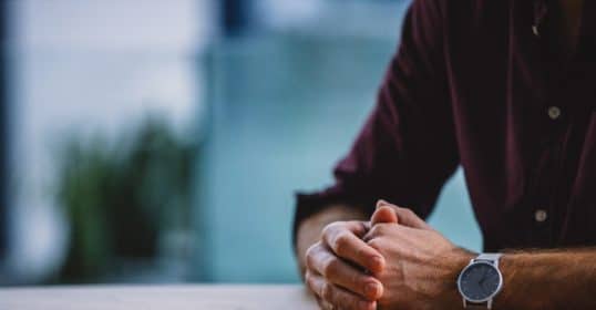 Close-up of a person's clasped hands resting on a table, waiting, wearing a watch on the left wrist.