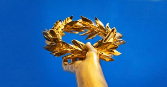 Golden laurel wreath, symbolizing attributes of greatness, held aloft against a blue sky.