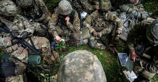 Soldiers in camouflage uniforms sitting on the ground during a briefing with maps and notepads for Podcast #892.