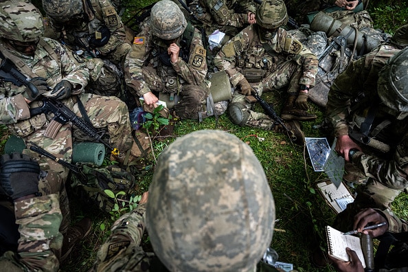 Soldiers in camouflage uniforms sitting on the ground during a briefing with maps and notepads for Podcast #892.