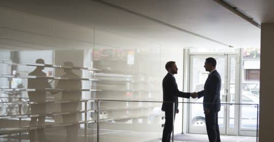 Two men shaking hands in a modern building entrance with reflective glass surfaces after discussing the Essential Guide on Podcast #899.