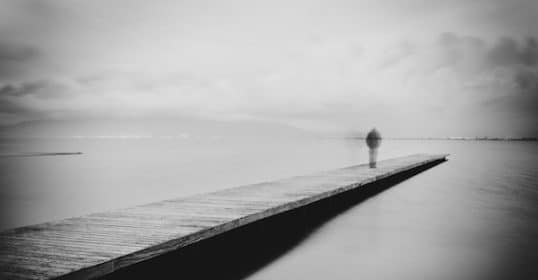 A solitary figure stands at the end of a pier, gazing out over a calm, hazy body of water, pondering suicide myths.