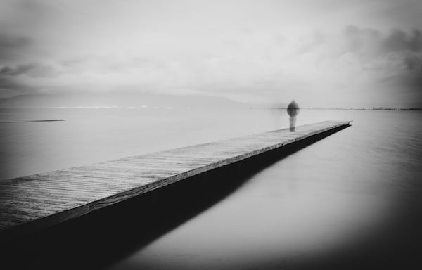 A solitary figure stands at the end of a pier, gazing out over a calm, hazy body of water, pondering suicide myths.