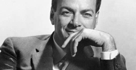 Black and white photo of a smiling man in a suit, resembling Richard Feynman, resting his chin on his hand.