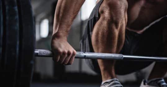 A person preparing to lift a barbell at a gym, focusing on hand grip and leg muscles, wonders how often to test their one-rep max.